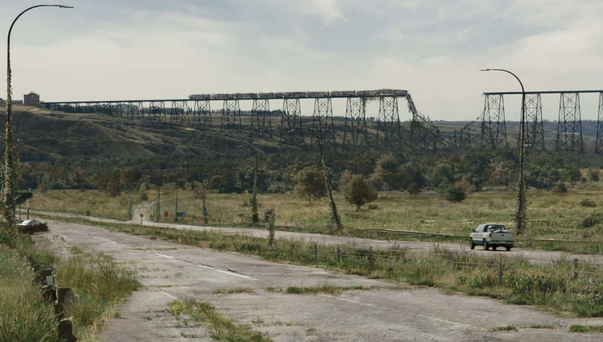 Blue and white truck drives down a decaying highway past a rail over bridge that has collapsed in the middle with a train on it from 'The Last of Us'